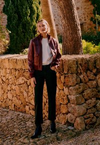 Femeie într-o jachetă de piele burgund, cămașă albă, pantaloni negri și cizme, stând cu o mână în buzunar lângă un zid de piatră, în aer liber.