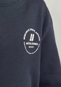 Jūros mėlynumo apvalios iškirpės marškinėliai, pagaminti iš medvilnės, su balta apvalia logotipą, kuriame užrašyta "JACK & JONES" su aplink jį esančiu tekstu.