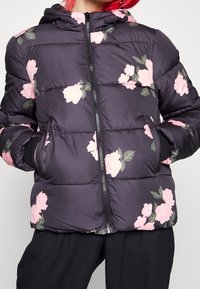 Černá prošívaná bunda s růžovým květinovým vzorem, vybavená předním zipem, kapucí a žebrovanými manžetami. Polstrovaná textura pro teplo a pohodlí.