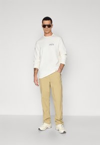 White long-sleeve shirt with a colorful logo, khaki pants, and white sneakers. Model wears oversized sunglasses. Simple, casual outfit.