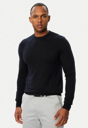 Homme portant un pull noir ajusté à manches longues et un pantalon gris clair, debout devant un fond blanc uni.