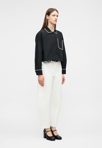 Mujer con camisa negra de manga larga con borde de perlas blancas, pantalones blancos de tiro alto y pierna ancha, y zapatos negros de tacón grueso.