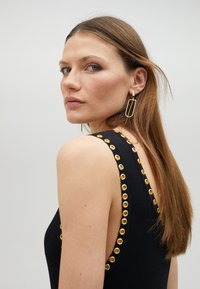 Černý bezrukávový top s pozlacenými dírkovými detaily na výstřihu a ramenou. Modelka má rovné vlasy a nosí geometrické náušnice.