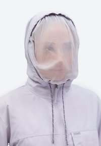 Άτομο που φοράει ανοιχτό μωβ μπουφάν με κουκούλα και λεπτό δίχτυ που καλύπτει το πρόσωπο μέσα στην κουκούλα.