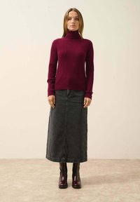 Burgundy ribbed turtleneck neule paita yhdistettynä pitkään mustaan farkkuhameeseen ja kiiltäväpintaisten burgundy nilkkasaappaisiin neutraalilla taustalla.