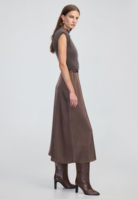 Brązowy bezrękawnik z dzianiny w połączeniu z długą, zwiewną brązową satynową spódnicą oraz brązowymi botkami na obcasie do kolan, tworząc elegancką sylwetkę.