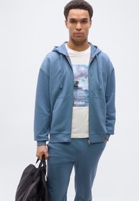 Νεαρός άνδρας με μπλε φούτερ με φερμουάρ και παντελόνι, κρατώντας μαύρη τσάντα, φοράει λευκό κοντομάνικο T-shirt με σχέδιο νερού και κείμενο "HUGO".