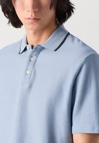 Hellblaues Poloshirt aus strukturierter Stoffqualität, mit einem Strickkragen in dunklen Marineakzenten, drei Knöpfen und kurzen Ärmeln.