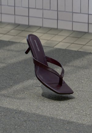 Donkerbruine sandaal met hak en vierkante neus, met dunne bandjes, zwevend boven een gestructureerde stoep nabij een betegelde muur.