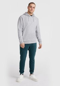 Sweat à capuche gris avec cordons de serrage ajustables, poignets et ourlet côtelés. Associé à un jogging teal et des baskets blanches. Design simple, sans motifs.