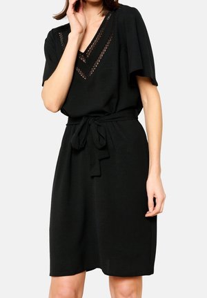 Femme portant une robe noire mi-longue à manches courtes, avec un col en V en dentelle et une ceinture en tissu nouée à la taille.