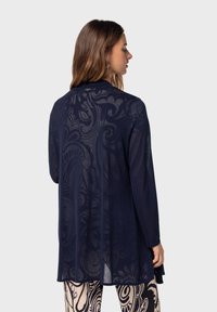 Marinblå genomskinlig cardigan med blommönster, långa ärmar och avslappnad passform, bärs över mönstrade byxor, med rund halsringning.