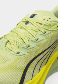 Közeli fotó egy sárga futócipőről, hálós felsőrésszel, fényvisszaverő fűzőrészletekkel és sötét csíkkal a világos sárga talpon.