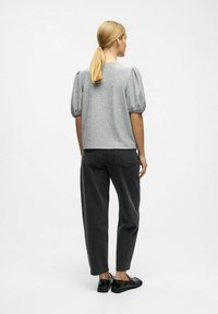 Kvinde med blond hestehale står med ryggen til iført grå kort puffærmet top, sorte tætsiddende jeans og sorte loafers på hvid baggrund.