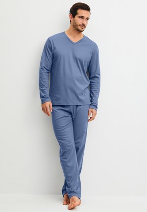 Man draagt een blauwe lange mouwen V-hals top en bijpassende broek, staat blootsvoets tegen een effen witte muur.