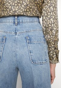 Jean bleu taille haute avec grandes poches arrière carrées, porté avec un chemisier à manches longues noir et beige à imprimé floral.