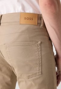 Beige chinos met een leren label "BOSS" op de tailleband, voorzien van standaard achterzakken en een gladde, gestructureerde stof.