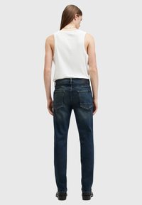 Jeans de mezclilla azul oscuro con corte recto, que cuentan con bolsillos traseros y una etiqueta de piel negra en la cintura, combinados con una camiseta sin mangas blanca.