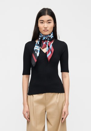 Γυναίκα με μακριά μαύρα μαλλιά, ντυμένη με μαύρο εφαρμοστό τοπ, μπεζ παντελόνι και πολύχρωμο μεταξωτό μαντήλι δεμένο γύρω από τον λαιμό της, πάνω σε απλό φόντο.