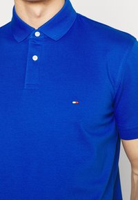 Blå polo t-shirt i strukturerad bomull med klassisk krage, som har tre vita knappar och en liten broderad logotyp i röd, vit och blå.