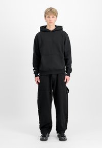 Jonge man staat rechtop en kijkt recht vooruit, gekleed in een zwarte hoodie, loszittende zwarte broek en zwarte sneakers, tegen een effen witte achtergrond.