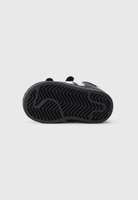 Semelle de sneaker en caoutchouc noir avec un motif de semelle ondulée, agrémentée d'une bande d'accent blanche. Le design présente des bords arrondis et une surface texturée.