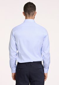 Camicia azzurra a maniche lunghe con colletto a punta, polsini con bottoni e tessuto liscio, indossata con pantaloni scuri su misura.
