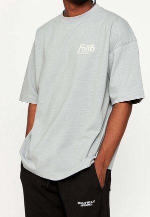 Mężczyzna ubrany w jasnoszarą, luźną koszulkę z krótkim rękawem z napisem „Faith” oraz czarne spodnie z logo „Multiply Apparel”, ręce w kieszeniach.
