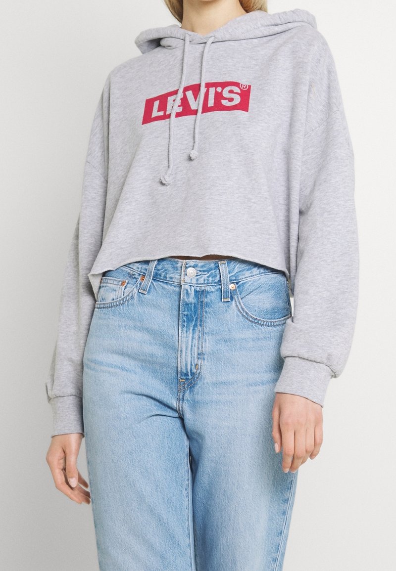 Personne portant un sweat à capuche court Levi's gris clair avec un logo rouge et un jean taille haute bleu clair, debout devant un fond uni.