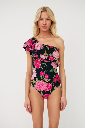 Femme portant un maillot de bain noir asymétrique avec un imprimé floral rose et rouge et un détail à volants, debout sur un fond blanc uni.