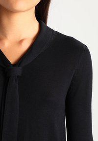 Granatowy sweter z dekoltem w serek i wiązaniem przy kołnierzu. Posiada długie rękawy i fakturowany materiał, prezentujący jednolity kolor.