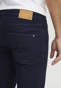 Jeans de mezclilla azul oscuro con un parche de cuero marrón etiquetado "ISOLID" en la cintura, que presenta un bolsillo trasero con un detalle de hardware mínimo.