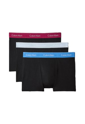 Spierbundige man met een donkere huid staat op blote voeten, gekleed in zwarte boxershorts met een witte tailleband tegen een effen grijze achtergrond.