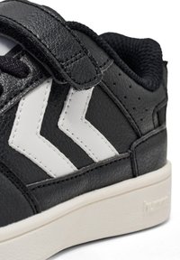 Schwarze Sneaker mit weißen Chevron-Streifen, ausgestattet mit einem Klettverschluss und einer strukturierten Lederoberfläche, die einen Nahaufnahme des oberen Abschnitts zeigt.