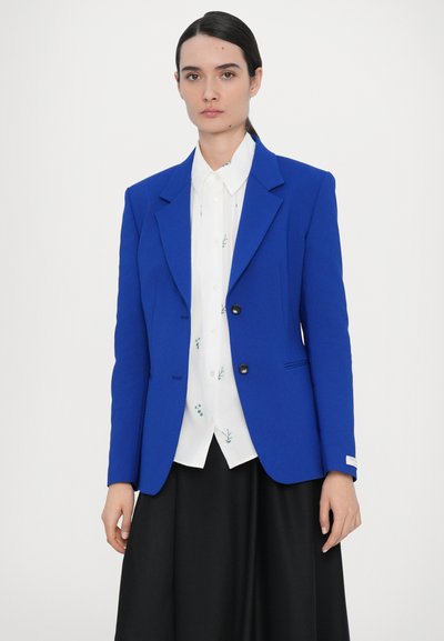 Blue structured blazer with two buttons, notch lapels, and side pockets, layered over a white shirt with green floral patterns. Black skirt.