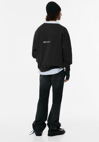 Černá oversized mikina s logem na zádech, přes kterou je nasazená světle modrá pruhovaná košile, doplněná černými džíny a černými botami.