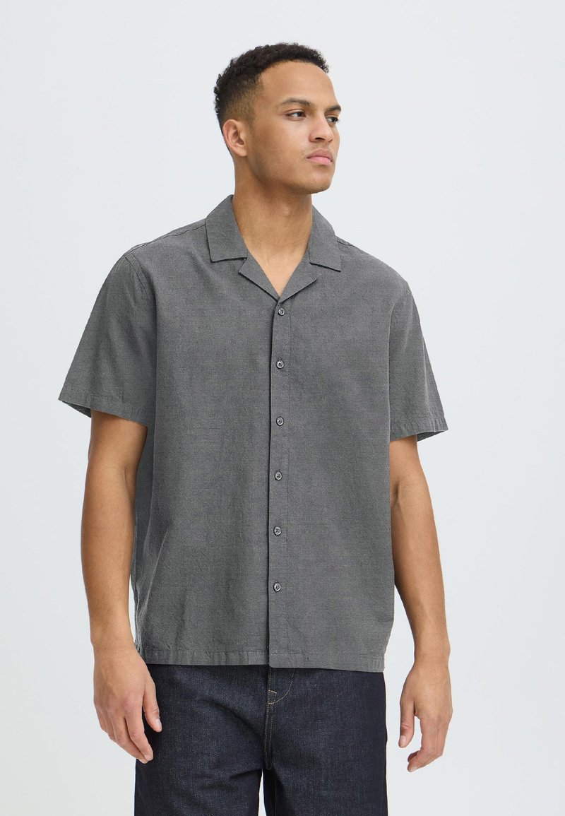 Homme portant une chemise grise à manches courtes boutonnée et un jean foncé, regardant sur le côté sur un fond clair uni.