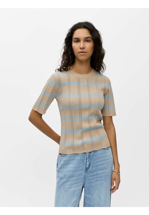 Kvinde med krøllet mørkt hår iført beige kortærmet striktop med blå striber og lyseblå jeans, stående mod en ensfarvet hvid baggrund.