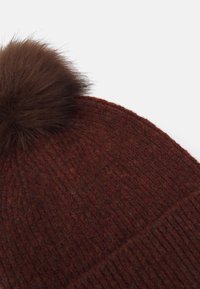 Gorro de lana en rojo oscuro con un acabado texturizado, que presenta un pom-pom de piel marrón en la parte superior. Diseño sencillo acanalado a lo largo del borde.