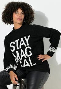 Suéter negro con texto en blanco y negrita "STAY MAGICAL", con cuello acanalado y detalles blancos semejantes a llamas en los puños. Combina con pantalones de cuero negros.