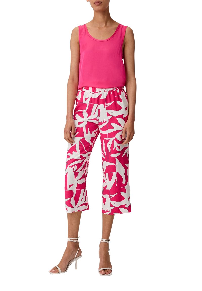 Femme portant un haut sans manches rose vif, un pantalon court rose et blanc à motif feuilles, et des sandales blanches à talons hauts avec brides.