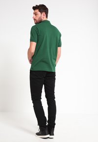 Camisa polo verde con dobladillo recto, con mangas cortas y cuello, combinada con pantalones negros y zapatos negros, vista desde la parte trasera.