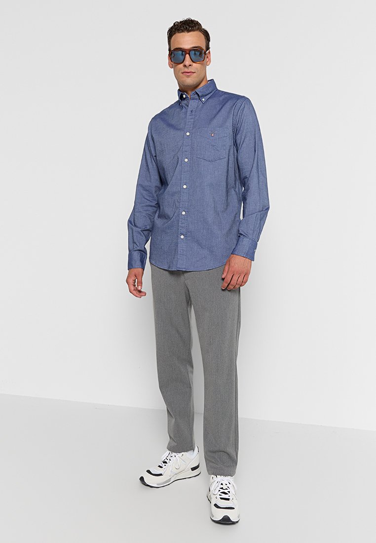 Camisa de botones de chambray azul con un bolsillo en el pecho, combinada con pantalones grises claros y zapatillas blancas con detalles en negro. Gafas de sol puestas.