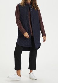 Gilet senza maniche trapuntato blu navy con bottoni a pressione, indossato sopra un maglione a maglia bordeaux, abbinato a pantaloni neri e scarpe da ginnastica bianche.
