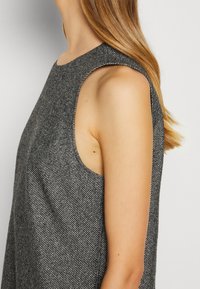 Femme aux longs cheveux bruns portant un haut sans manches texturé gris foncé, vue de côté avec un focus sur l'épaule et le haut du bras.