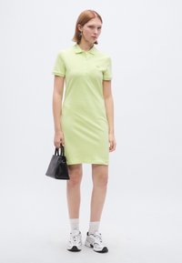 Robe polo vert clair à manches courtes, avec col et boutons. Associée à un petit sac à main noir et à des baskets blanches avec des accents gris.