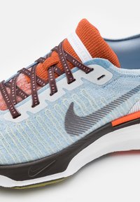 Světle modrá běžecká obuv s texturovanou tkaninou, oranžovými a burgundskými tkaničkami a černou podrážkou. Obsahuje výrazné černé logo Nike swoosh.