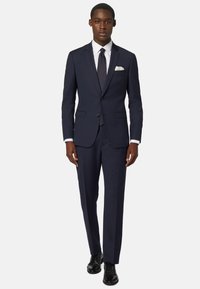 Marineblauer Anzug mit taillierter Passform, leichtem Stoff, zwei Knöpfen, Reverskragen und weißem Taschentuch. Schwarze Dressschuhe runden das Outfit ab.