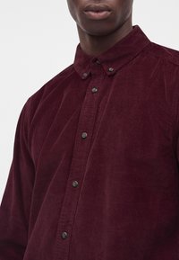 Gros plan d'une personne portant une chemise en velours côtelé bordeaux avec un col boutonné sur un fond blanc.