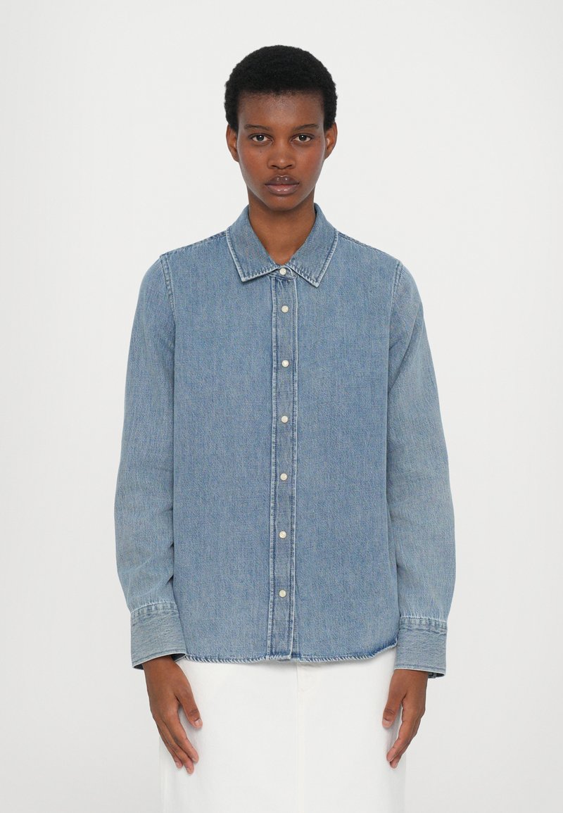 Light blue denim shirt with long sleeves, button-down front, pointed collar, and subtle stitching details. Soft texture with a relaxed fit.